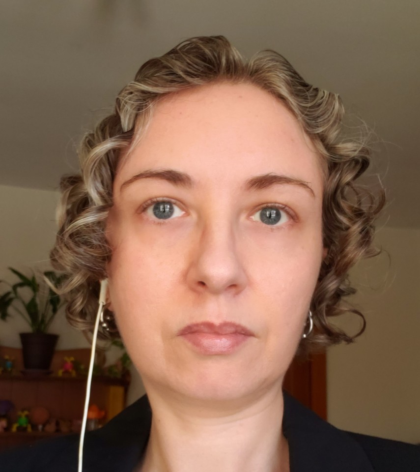 A headshot of Dawn Lynch, a white nonbinary person with short wavy hair and blue eyes. They are looking at the camera with a serious expression, wearing a black blazer. There is one wired earbud in their right ear and silver hoop earrings in both ears. In the background there's a shelf with knickknacks and plants.