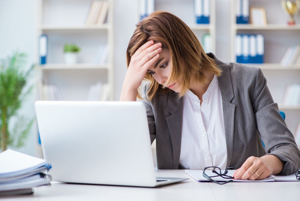 A businesswoman sits at a laptop with her hand on her forehead.